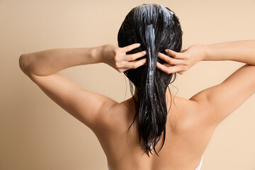 Young woman applying hair mask on beige background, back view