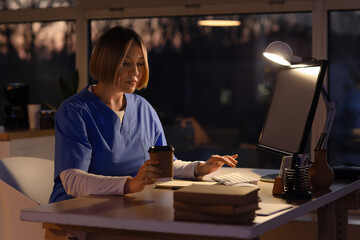 Fototapeta na wymiar Female nurse with coffee cup working evening shift at table in clinic