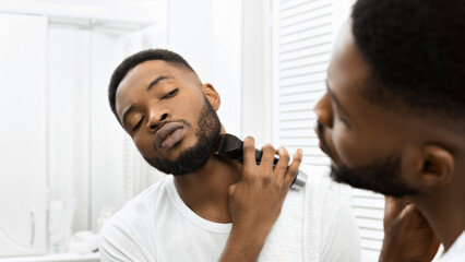Man trimming beard with electric clippers in mirror in bathroom - obrazy, fototapety, plakaty