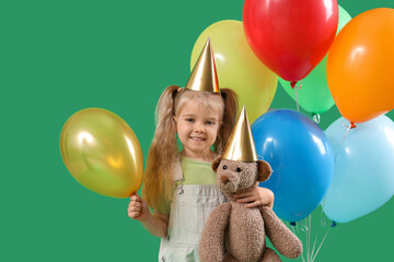 Happy smiling little girl in birthday hat with colorful balloons and toy bear on green background