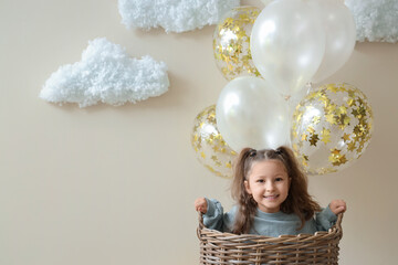 Cute little girl in basket with balloons and cotton clouds on light background
