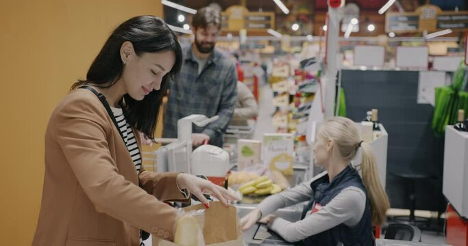 Young woman buying food in supermarket packing purchase at counter while cashier working scanning groceries. Shopping and retail business concept.
