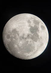 A high-resolution picture of the bright full moon shines in the dark sky, revealing its bumpy surface adorned with dark spots and bright spots reflecting light.