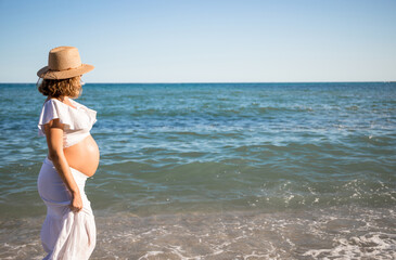 Pregnant woman with sun hat during her walk on the beach relaxing on her summer vacation, happy pregnancy outdoors.