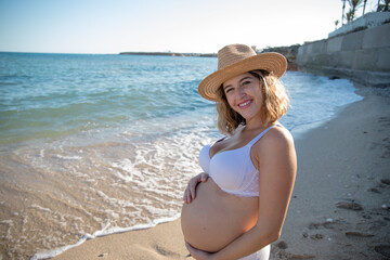 Pregnant woman with sun hat during her walk on the beach relaxing on her summer vacation, happy pregnancy outdoors.