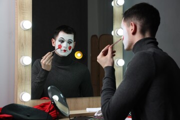 Young man applying mime makeup near mirror in dressing room - Powered by Adobe
