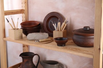 Set of different crafting tools and clay dishes on wooden rack in workshop