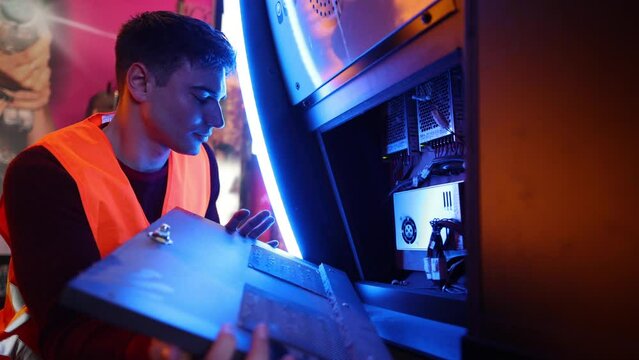  Concentrated arcade technician in high-visibility vest examining the internal components of a video game machine. Arcade Maintenance Technician Inspecting Interior of Game Cabinet