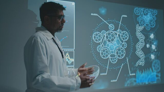 Medium footage of mature Asian male physician standing next to projector screen at meeting, speaking about scientific research on biochemistry of new drug, pointing to slide with cell and molecules