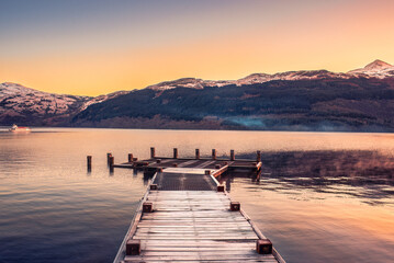 A pier extending over the calm waters of Loch Lomond at sunrise in Scotland