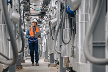 man in a safety vest is walking through a large industrial building with pipes and wires. He is holding a clipboard inspecting pipeline area.