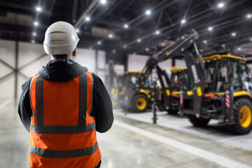 Builder in hangar with construction equipment. Man working with his back to camera. Machines for construction and engineering work. Construction worker in reflective vest. Builder chooses bulldozer - obrazy, fototapety, plakaty