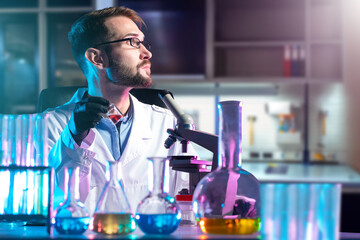 Obraz na płótnie Canvas Man microbiologist. Laboratory assistant with microscope and flasks. Multi-colored reagent on microbiologist table. Scientist sits at laboratory desk. Microbiologist looks away. Man biologist at work
