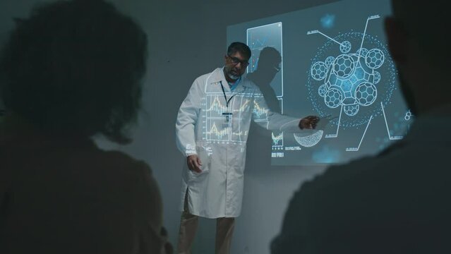 Wide shot of mature Asian male researcher standing on stage next to projector screen, speaking about new scientific discovery in immunology, pointing on slide with cell drawings, talking to public