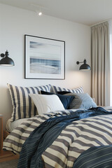 A modern bedroom with striped bedding in navy blue and white stripes, black sconces on the wall above the bed, a framed print of an ocean scene hanging over the bedside table, and a wooden nightstand.