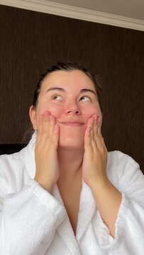 Vertical video. Young adult woman in white bath robe applies moisturizer to face in hotel room. Concept of rest, skin protection during cold season, cosmetic procedure, spa salon, relax, march 8	