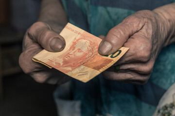 Underpaid job in Brazil, Concept, Old woman holding 20 reais banknote in her worn hand, Gloomy dark colors - 772231781