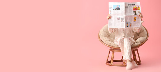 Young woman reading newspaper in armchair on pink background