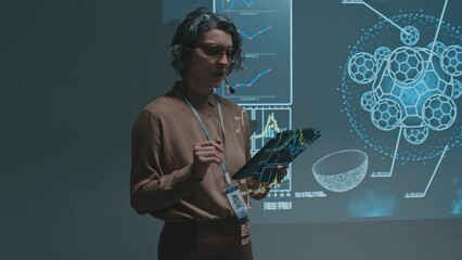 Medium shot of Caucasian female scientist or professor with tablet computer and headset microphone giving lecture on biochemistry at university or conference