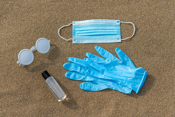 Summer beach layout with sunglasses, protective gloves, medical mask and hand sanitizer on sand....