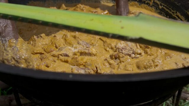 A man adds spices and stirs them together with pieces of meat in a large iron pot