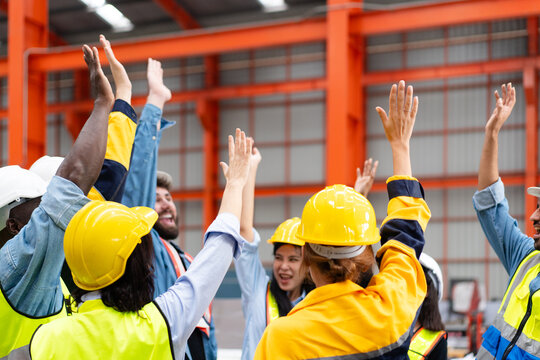 group of diverse workers raising hands to celebrate project success at manufacturing industrial factory, team colleagues in safety clothes smile glad shouting about production progress in the meeting