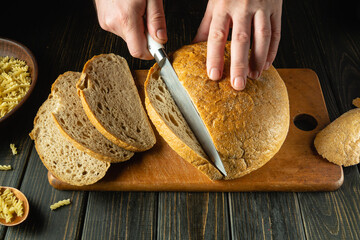 A cook cuts fresh bread with a knife on a kitchen board close-up. Slicing rye bread on the kitchen table or concept of healthy eating and traditional baking