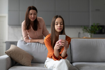 Parental monitoring of teens. Curious mother spying daughter, girl messaging on smartphone on sofa...