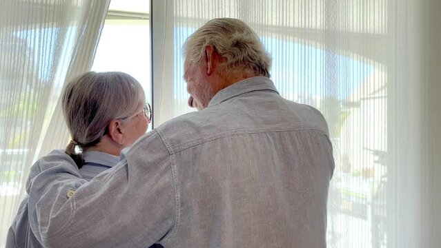 Smiling bonding senior caucasian couple looking out the window, senior man and woman supporting each other in a time of melancholy or trouble