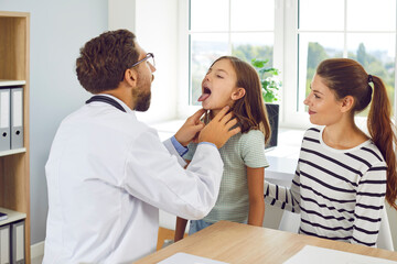 Male family pediatrician examining little girl patient's throat in medical clinic during checkup. Doctor otolaryngologist exam the oral cavity of a sick child sitting with her mother in the office.