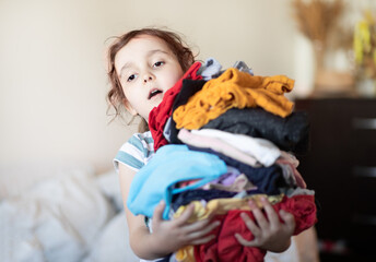 Funny cute girl kid holding messy stack pile of clothes things. Home chores housework. Child...