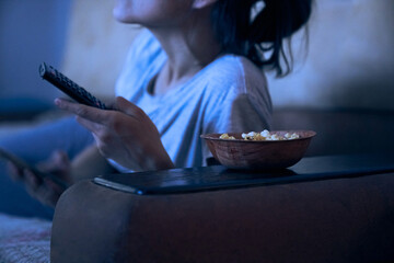 Close up view of woman watching TV at night, lying on sofa with plate, holding remote control....