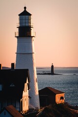 Idyllic scene of a lighthouse by the ocean at sunset, silhouettes of nearby buildings in background © Wirestock