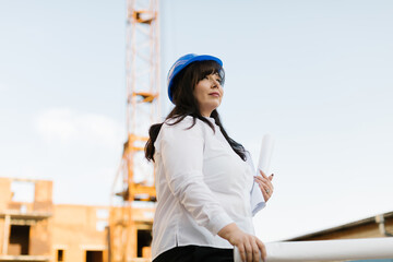 Plus size woman engineer in a white shirt and blue helmet is holding blueprints. Female in blue helmet architect is holding blueprints at a construction site.