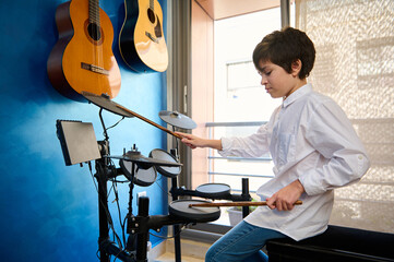 Teenage boy in white shirt and blue jeans, playing drum kit in his modern music studio room. Two...