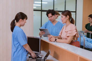 Asian female administrator talks with patient. Adult woman stands near reception desk in clinic lobby area, asks information, makes appointment with doctor. Medical staff work in modern hospital.