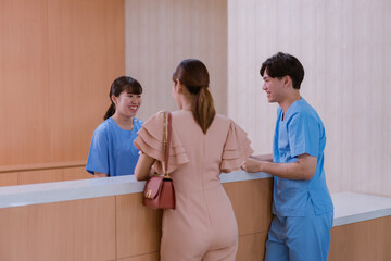 Asian female administrator talks with patient. Adult woman stands near reception desk in clinic lobby area, asks information, makes appointment with doctor. Medical staff work in modern hospital.