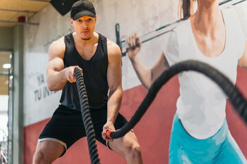 Muscular redhead man in tank top vigorously moving ropes in workout.