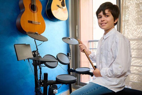 Teenager boy drummer musician in white shirt and blue jeans, sitting at drum set in his retro music studio , holding drumstick and smiling looking confidently at camera. Guitars hanging on a blue wall