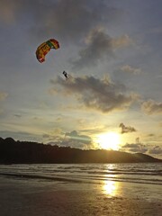 The tranquil ocean at sunset, framed by a dark mountain silhouette, gentle light peeking through the clouds, and a paragliding adventure in the distance.