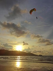 The tranquil ocean at sunset, framed by a dark mountain silhouette, gentle light peeking through the clouds, and a paragliding adventure in the distance.