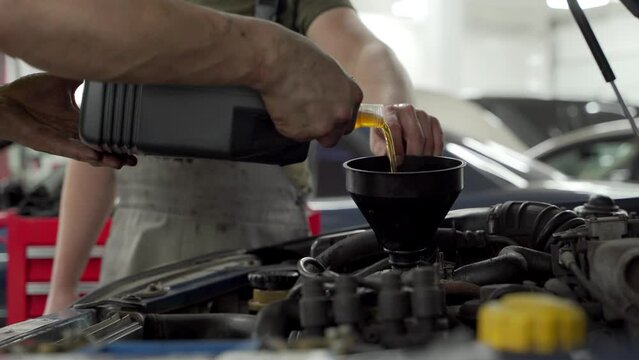 Auto mechanic pours fresh motor oil into engine using funnel at service station. Vehicle maintenance, oil change process in garage. Automotive technician performs fluid replacement, car care.