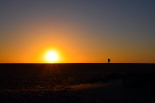 Against the background of the setting sun in the desert, silhouettes of two travelers in the distance.