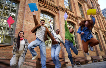 Happy group of multiethnic students celebrating the last day of school, looking at camera and jumping raising arms holding university workbooks outside campus.