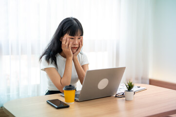 Worried Asian woman looking at laptop screen, expressing stress and concern with smartphone and...
