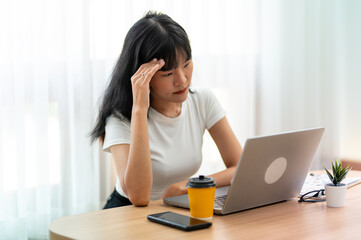 Worried Asian woman looking at laptop screen, expressing stress and concern with smartphone and...