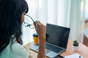 Asian woman wearing glasses works seriously on laptop in bright room. - obrazy, fototapety, plakaty