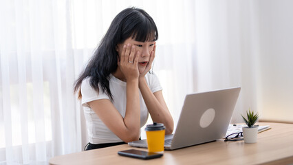 Worried Asian woman looking at laptop screen, expressing stress and concern with smartphone and coffee on desk.