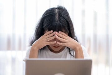 Worried Asian woman looking at laptop screen, expressing stress and concern with smartphone and...