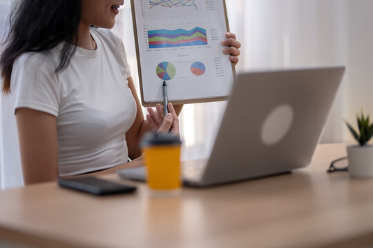 Crop shot of asian woman presents a clipboard with business data analysis charts during an online meeting via laptop.
